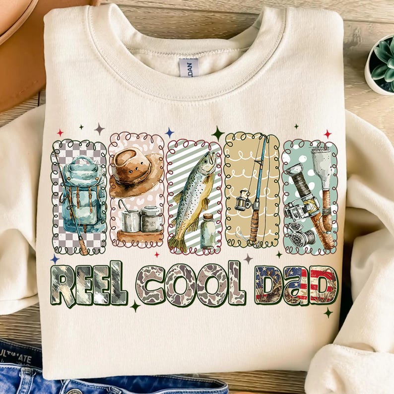 May include: Cream-colored sweatshirt with a fishing theme. The design includes illustrations of fishing gear, a fish, and the text "REEL COOL DAD" in a mix of fonts and patterns. The overall style is casual and outdoorsy.
