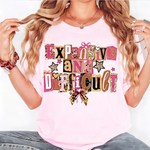 May include: A pale pink t-shirt featuring the text "Expensive and Difficult" in a variety of fonts and colours, including pink, brown, and leopard print. The design incorporates stars and bows.