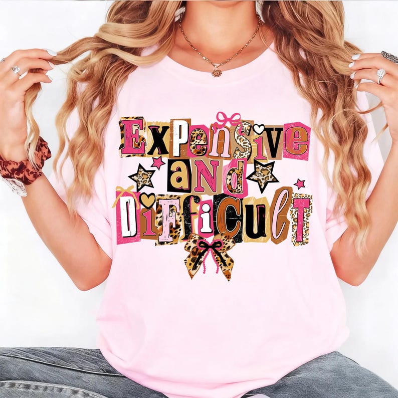 May include: A pale pink t-shirt featuring the text "Expensive and Difficult" in a variety of fonts and colours, including pink, brown, and leopard print. The design incorporates stars and bows.