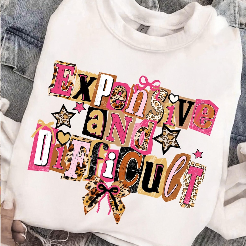 May include: White sweatshirt featuring the phrase "Expensive and Difficult" in a collage design. The text incorporates pink, brown, black, and leopard print patterns, with star and bow embellishments.