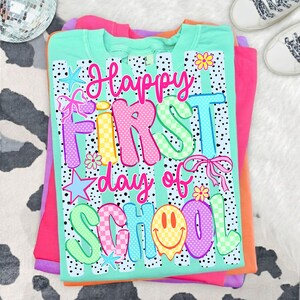 May include: A mint green t-shirt featuring a black and white polka dot pattern and the colorful text Happy First day of SCHOOL. The design includes graphic elements such as pink bows, yellow flowers, blue stars, and a yellow smiley face.