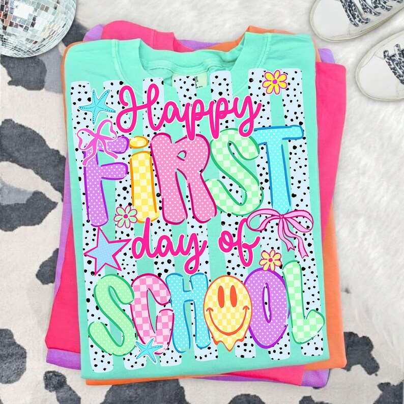 May include: A mint green t-shirt featuring a black and white polka dot pattern and the colorful text Happy First day of SCHOOL. The design includes graphic elements such as pink bows, yellow flowers, blue stars, and a yellow smiley face.