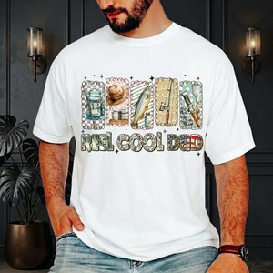 May include: White t-shirt with a colorful graphic featuring fishing-related images and the text "REEL COOL DAD." The graphic includes a backpack, hat, fish, fishing rod, and other fishing gear. The text is in a camouflage pattern.