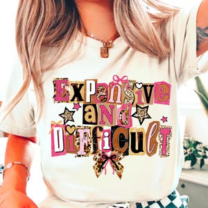 May include: A cream-coloured t-shirt featuring the phrase "Expensive and Difficult" in a collage design. The text incorporates pink, brown, and leopard print patterns, with star and bow accents.