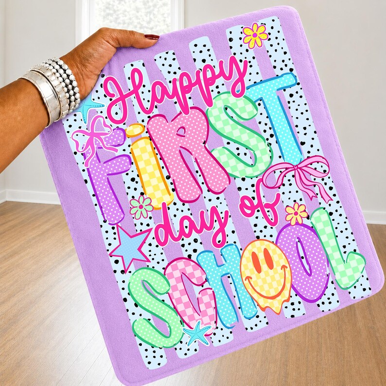 May include: A rectangular lavender fabric sign featuring the text Happy First day of School in colorful, patterned bubble letters. The design includes black polka dots, vertical stripes, pink bows, yellow flowers, blue stars, and a yellow smiley face.