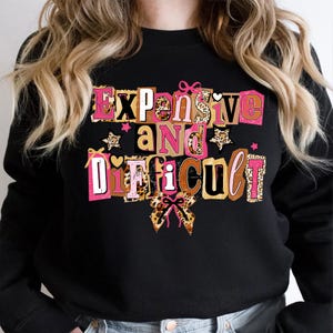 May include: Black sweatshirt featuring the phrase "Expensive and Difficult" in pink, brown, and leopard print lettering. The design includes stars and a bow. A casual top for everyday wear.