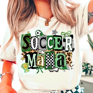 May include: Off-white t-shirt featuring the text "Soccer Mama" in a green, black, and white design. The design includes soccer balls, stars, and a leopard print pattern. A trophy and football boot are also featured.