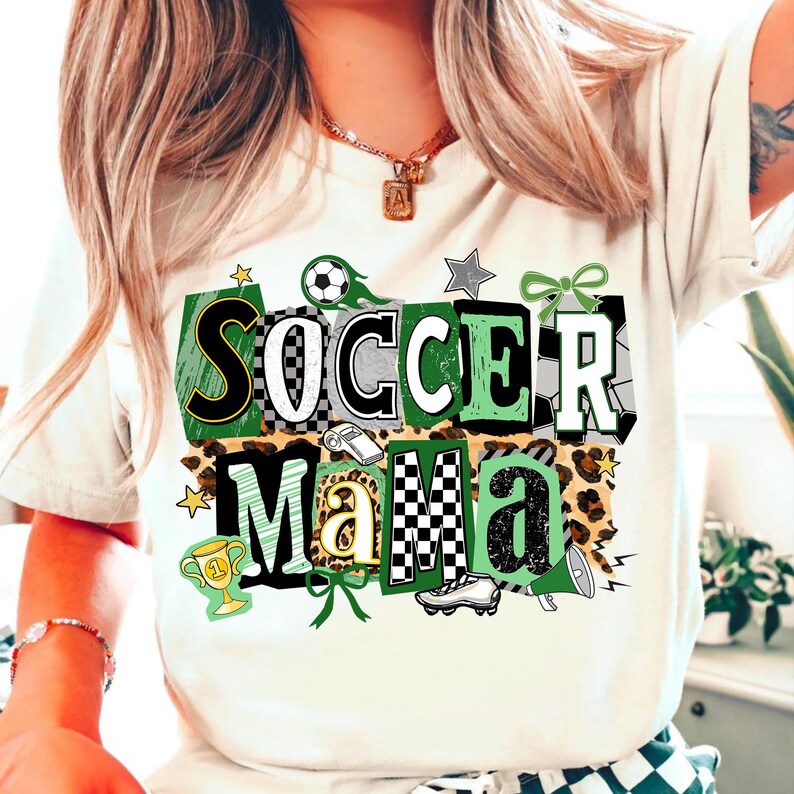 May include: Off-white t-shirt featuring the text "Soccer Mama" in a green, black, and white design. The design includes soccer balls, stars, and a leopard print pattern. A trophy and football boot are also featured.