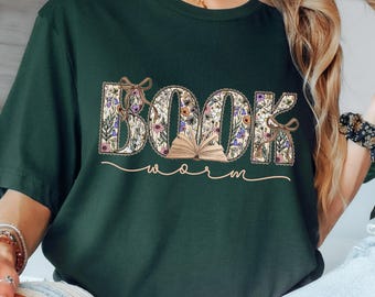 May include: Dark green t-shirt with the words "BOOK worm" in a floral design. The letters are filled with flowers and leaves, and the word "worm" is written in a cursive font. The shirt is a casual style.