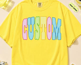 May include: A bright yellow short-sleeve t-shirt featuring the word CUSTOM printed in large, multi-colored, vertically striped block letters across the chest. The shirt has a crew neckline and a Comfort Colors brand label at the inner collar.