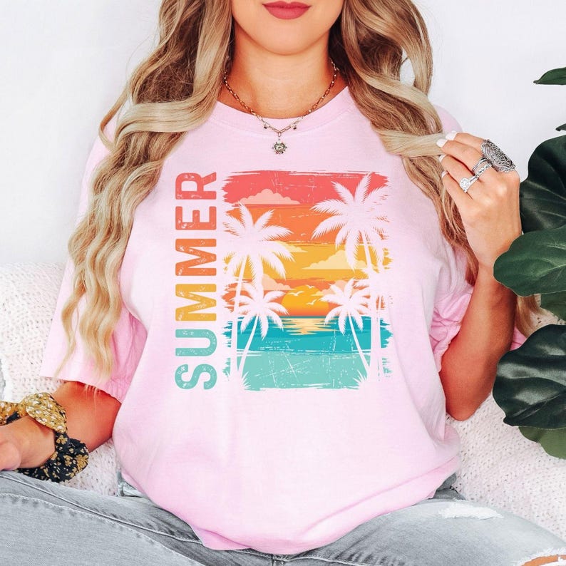 May include: A pale pink t-shirt with the word "SUMMER" printed vertically in colourful letters. The shirt features a graphic of palm trees, a sunset, and ocean waves.