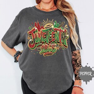 A person wearing a grey t-shirt with a colorful graphic design that says "JUNEEENTH 1865" and includes various decorative elements such as stars, flowers, and a city skyline.