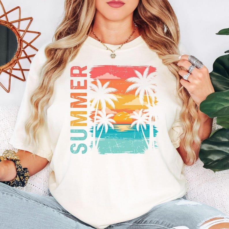 May include: Cream-coloured t-shirt with a vibrant graphic of a tropical sunset, palm trees, and the word "SUMMER" in vertical text. The design incorporates red, orange, yellow, and blue hues, creating a summery atmosphere.