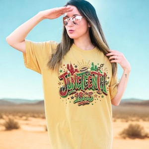A woman wearing a yellow t-shirt with a graphic design and text, standing in a desert landscape with mountains in the background.