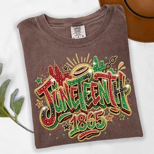 A brown t-shirt with a colorful graphic design featuring the text "JUNEENTH 1865" and decorative elements such as stars and lightning bolts.