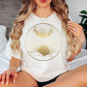 Może przedstawiać: Kremowy t-shirt z złotym niebiańskim wzorem. Grafika zawiera słońce, księżyc i pasmo g&oacute;rskie w okrągłej ramie. Projekt jest wyśrodkowany na przodzie koszulki, tworząc minimalistyczną i stylową estetykę.