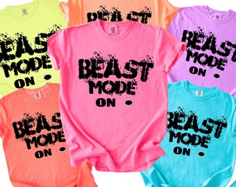 May include: A collection of six short-sleeved crew neck t-shirts in neon yellow, orange, pink, purple, coral, and turquoise. Each shirt features the black text BEAST MODE ON in a distressed, splattered font style centered on the chest area.