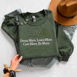 A green sweatshirt with a butterfly design and the text "Dream More. Learn More. Care More. Be More." is displayed on a bed, along with a hat and a pair of jeans. A hand is visible in the foreground, holding a bracelet.