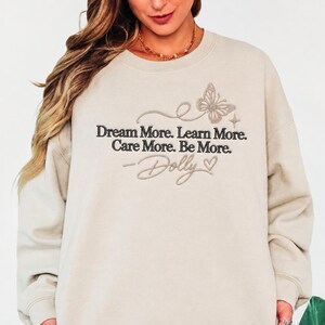 A woman wearing a beige sweatshirt with the text "Dream More. Learn More. Care More. Be More." and a butterfly design printed on it.