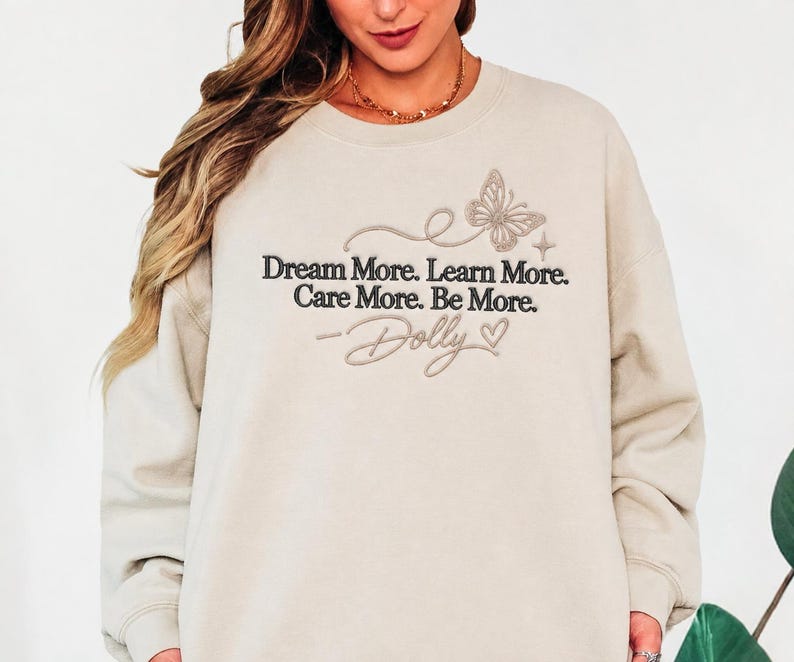 A woman wearing a beige sweatshirt with the text "Dream More. Learn More. Care More. Be More." and a butterfly design printed on it.