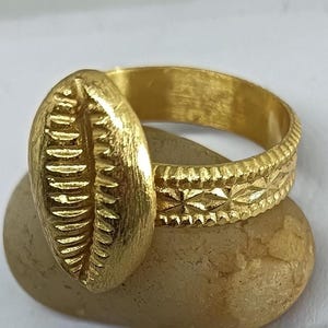 Puede incluir: Un anillo dorado con un diseño texturizado de concha de cauri. La banda tiene un patrón geométrico detallado. El anillo se muestra sobre una pequeña piedra de color claro, sobre un fondo blanco. Este anillo es una joya.