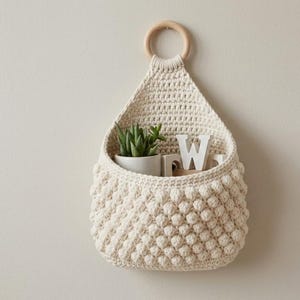 May include: Cream-colored, handmade crochet wall hanging with a wooden ring for hanging. The basket-like pouch is filled with a small potted plant and a white letter "W". The wall hanging is mounted on a neutral-colored wall.