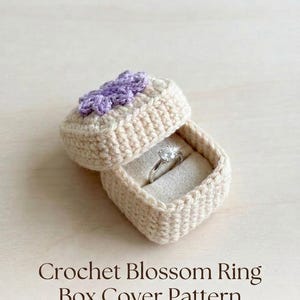 May include: A square crochet ring box in a light beige yarn featuring a small purple flower embellishment on the lid. The open box contains a silver ring with a clear stone. The text Crochet Blossom Ring Box Cover Pattern is below.