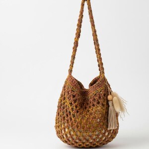 May include: A crocheted shoulder bag in shades of brown, yellow, and green. The bag has a net-like design and a long strap. A tassel with a wooden bead and dried flowers hangs from the side. The bag is designed to be carried over the shoulder.