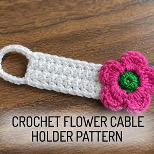 May include: A white crocheted cable holder featuring a pink flower with a green center at one end and a circular loop at the other. The text CROCHET FLOWER CABLE HOLDER PATTERN is printed in black below the item on the wooden surface.