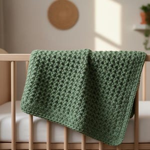May include: A handmade, green crochet baby blanket draped over a wooden cot. The blanket has a textured, woven appearance and is a soft, muted green colour. The cot has a white mattress and is made of light-coloured wood.