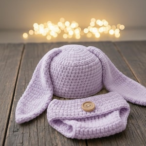 Könnte beinhalten: Ein helllila Häkel-Hasenmützen- und Windel-Set. Die Mütze hat lange, schlaffe Ohren. Die Windel hat einen Holzknopfverschluss. Das Set ist aus weichem Garn gefertigt und für ein Baby konzipiert.