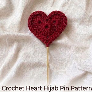 Op de afbeelding: Een donkerrood gehaakt hart bevestigd aan een lange, dunne, goudkleurige metalen speld. Het hart heeft een getextureerde, met de hand gestikte garenconstructie met een centraal punt. De tekst Crochet Heart Hijab Pin Pattern is onderaan zichtbaar.