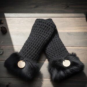 May include: A pair of black knitted arm warmers with a fur trim at the wrist. Each arm warmer has a small, round wooden tag attached. The arm warmers are lying on a dark wooden surface.