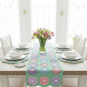 May include: A dining table set for a meal, featuring a crocheted floral table runner in green, pink, and purple. White plates and bowls are set on matching placemats and coasters. A vase of colorful flowers sits in the center.