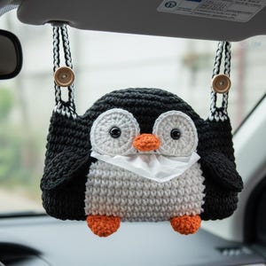 Puede incluir: Una funda de caja de pañuelos de ganchillo hecha a mano con forma de pingüino. El pingüino tiene cabeza y alas negras, cuerpo gris, ojos blancos y pico y patas naranjas. La funda de la caja de pañuelos cuelga de la visera del coche con botones de madera.