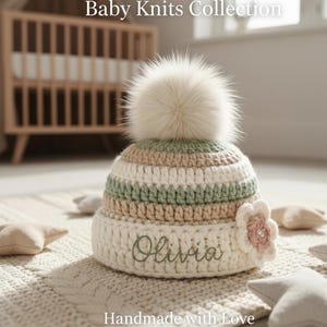 Puede incluir: Un gorro de punto para bebé de color crema con un gran pompón esponjoso de color blanquecino. El gorro presenta rayas horizontales en crema, verde claro y beige. El nombre "Olivia" está bordado en verde. Una flor crema con un centro rosa adorna el lateral. El texto "Baby Knits Collection" y "Handmade with Love" son visibles.
