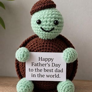 May include: A crocheted turtle figurine with a light green head and limbs, a dark brown shell, and a matching brown cap. The turtle holds a white rectangular card with the printed text: Happy Father’s Day to the best dad in the world.