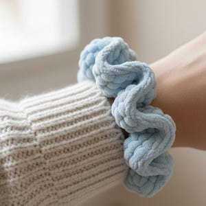 May include: A light blue, chunky knit scrunchie. The scrunchie is made of a soft, textured material and is wrapped around a wrist. The wrist is covered by a light gray, knitted sweater.
