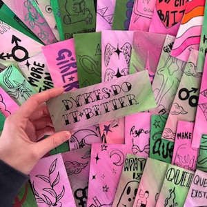 Op de afbeelding: Een verzameling kleurrijke, handgeschilderde bladwijzers met diverse ontwerpen en tekst. De bladwijzers bevatten illustraties van een kikker, engelen vleugels en de tekst "DYKES DO IT BETTER". De kleuren zijn roze, groen en wit.