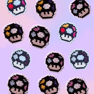 May include: A collection of twelve cross-stitch patches shaped like skulls. Each skull features a black base with a grid-like texture and is decorated with small, colourful pixelated accents in shades of yellow, pink, blue, and white.