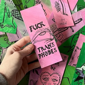 May include: A selection of handmade bookmarks in pink and green hues. One pink bookmark displays the text "FUCK TRANS PHOBES" with an eye drawing. Other bookmarks feature illustrations and text such as "pillow princess" and "lavender menace".