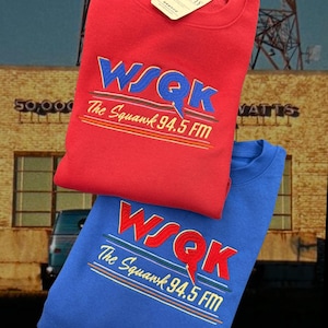 May include: Two crewneck sweatshirts, one red and one blue, featuring the embroidered text "WSQK The Squawk 94.5 FM" in blue and red. The red sweatshirt has a tag attached. The background shows a building and a radio tower.
