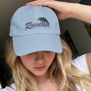 May include: Pale blue baseball cap featuring the text "Ravens Basketball" in purple script and a black raven graphic. The cap is made of a soft material and has a curved peak. A casual accessory for sports fans.
