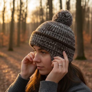 以下が含まれることがあります： 茶色と白のニットの冬用帽子で、ポンポンが付いています。帽子には耳当てと、下部に濃い茶色のバンドが付いています。画像内の人物はグレーのセーターを着ています。