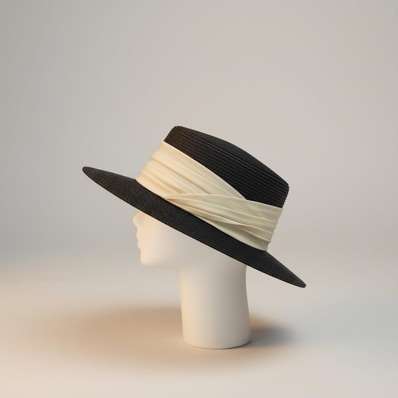 May include: A black fedora-style hat with a wide brim and a cream-colored band. The hat is displayed on a white mannequin head against a plain background. The hat's design is simple and elegant.