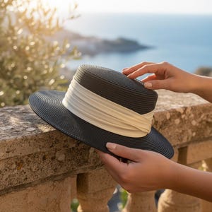 May include: A dark gray sun hat with a wide brim and a white band. The hat is held by a person on a stone wall overlooking the ocean. The hat's brim is approximately 12 inches in diameter.