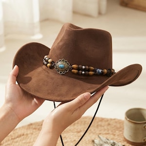 Könnte beinhalten: Ein brauner Filz-Cowboyhut mit einem dekorativen Band aus Holzperlen und Metallabstandshaltern. In der Mitte des Bandes befindet sich ein verziertes, gewelltes Metall-Concho-Element, das mit einem ovalen, türkisblauen Stein besetzt ist.