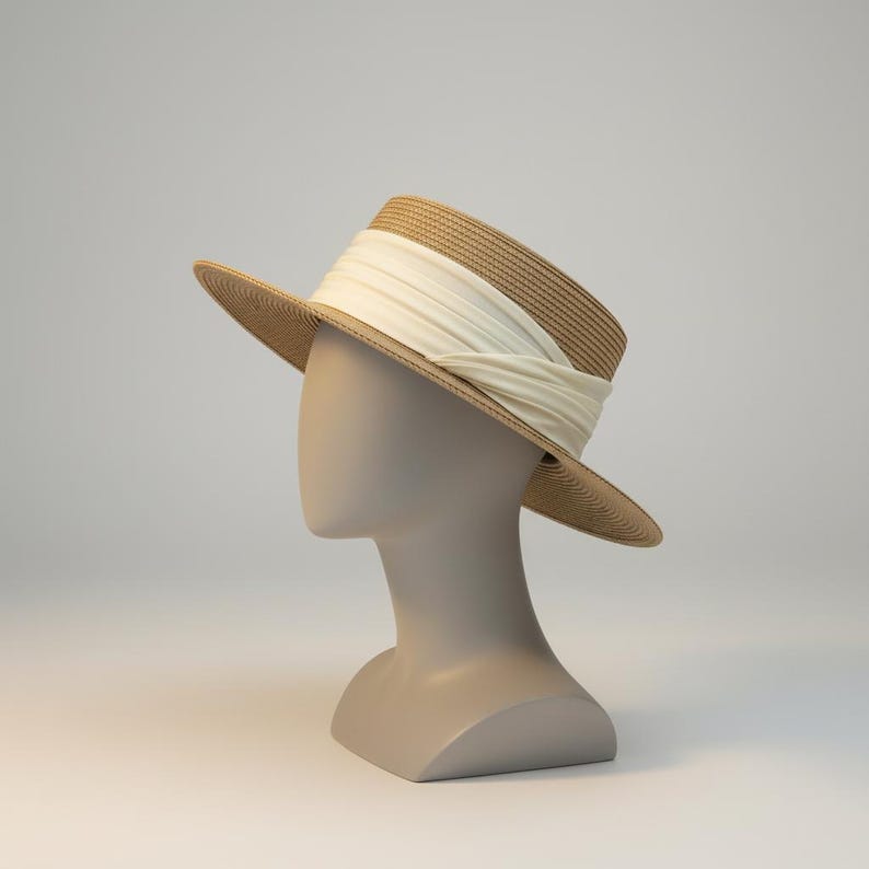 May include: A tan straw hat with a wide brim and a cream-colored ribbon wrapped around the crown. The hat is displayed on a mannequin head against a neutral background. The hat's design is classic and elegant.