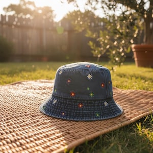 May include: A denim bucket hat with embroidered colorful flowers. The hat is sitting on a woven mat in a sunny outdoor setting. The hat's brim is a darker shade of denim.