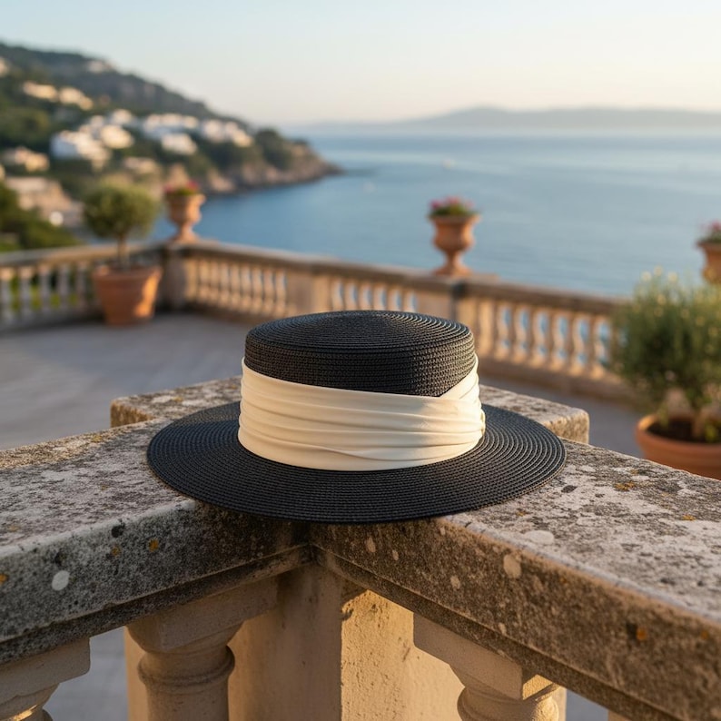 May include: A black sun hat with a wide brim and a cream-colored band. The hat is sitting on a stone ledge with a scenic ocean view in the background. The hat's design is classic and elegant.